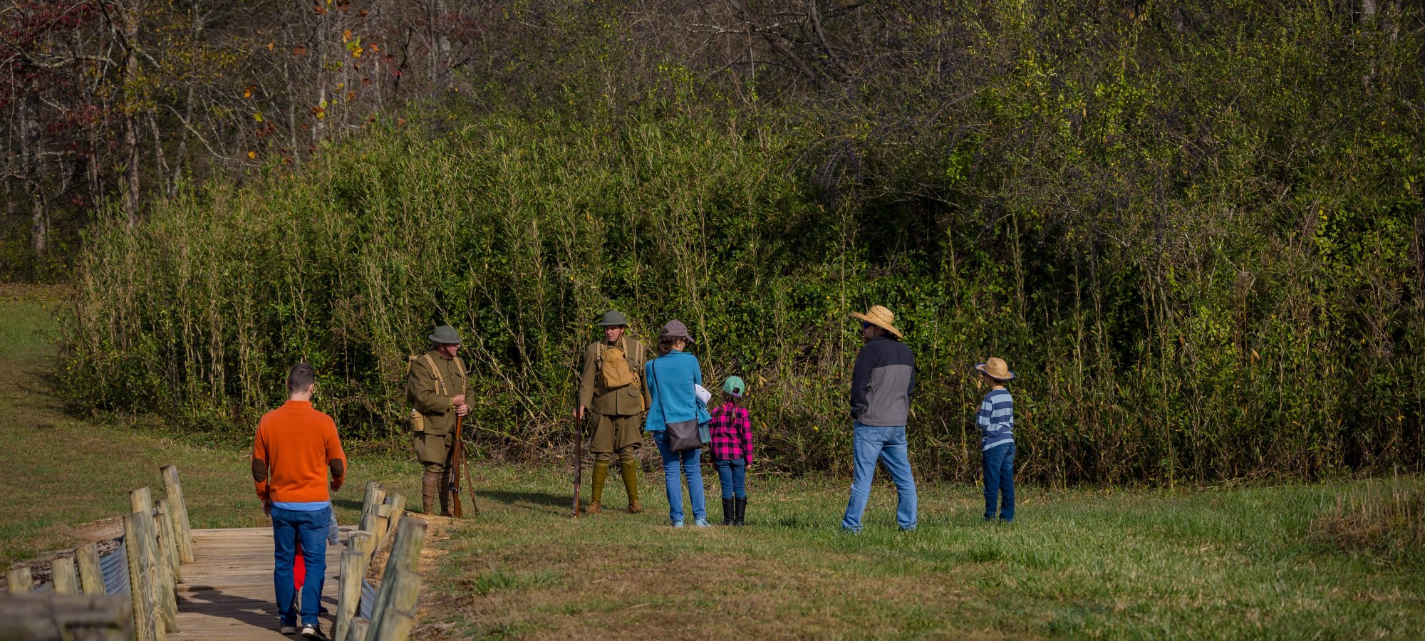 A group of people, including two in historical military uniforms, interact outdoors near a wooded area. The scene conveys a peaceful, educational vibe.