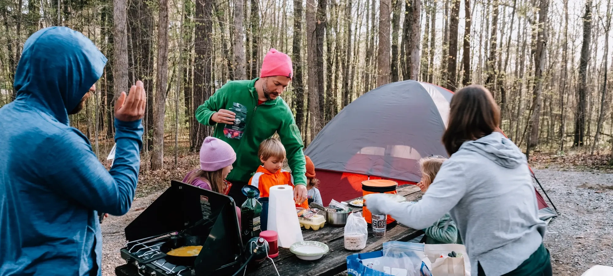 Family cooking at a campsite with a tent and trees in the background.
