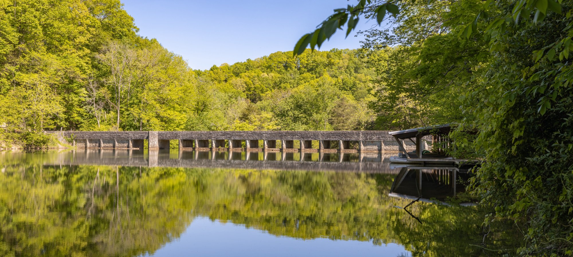 A serene scene with a stone bridge over a calm river, surrounded by lush green trees. The clear sky reflects on the water, creating a tranquil atmosphere.
