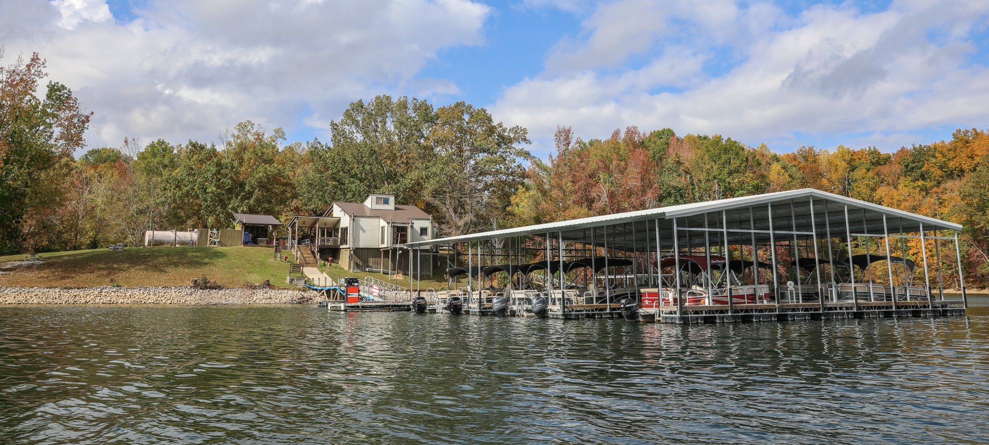 Lake scene with a boat dock and modern house in lush autumnal trees. Bright blue sky with clouds, creating a serene and peaceful atmosphere.