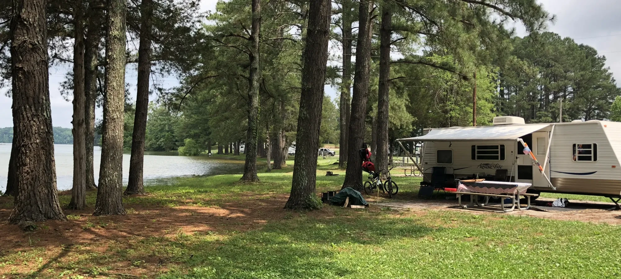 Camper by a lake surrounded by tall pine trees, with picnic table and bicycles nearby.