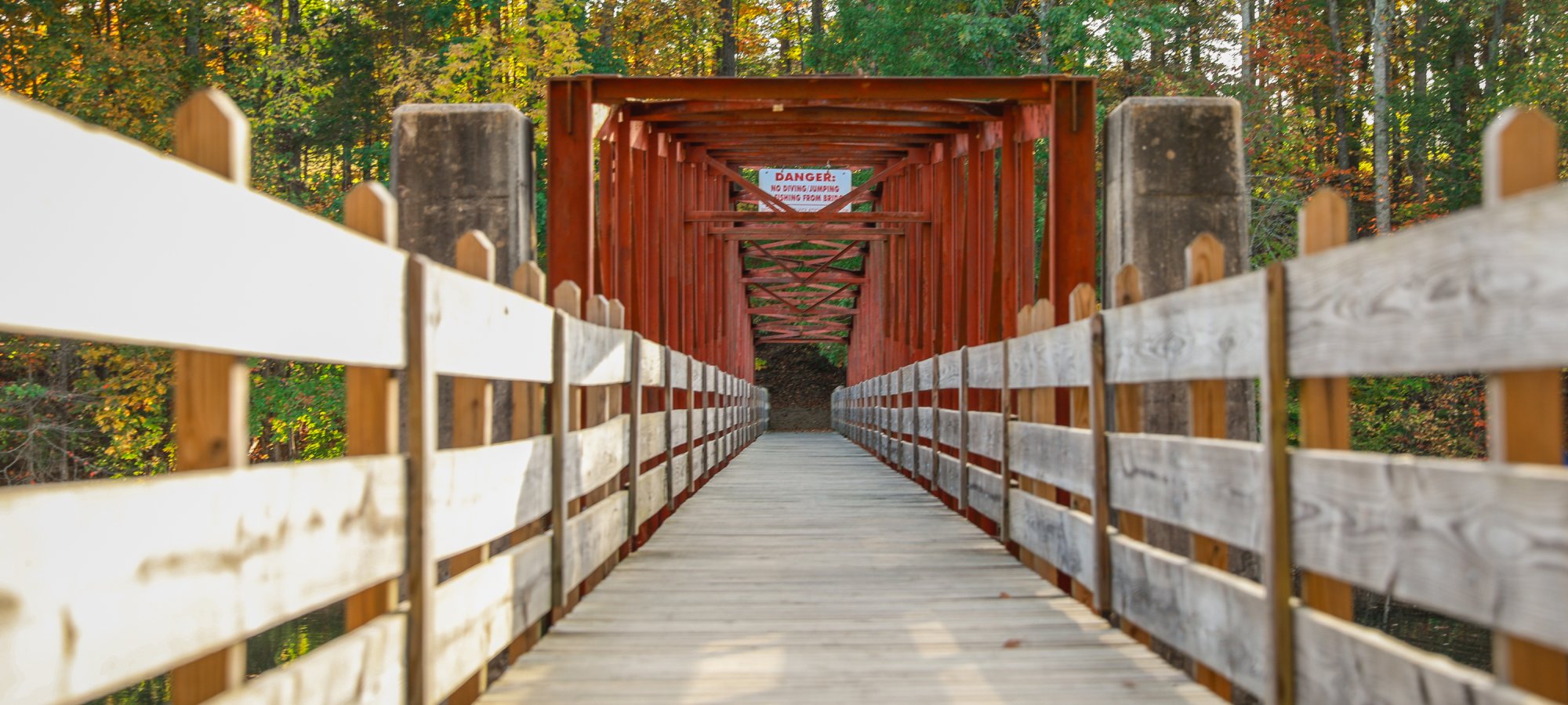 A wooden bridge with red beams extends through a leafy forest in autumn. Sunlight casts soft shadows, creating a serene and inviting atmosphere.