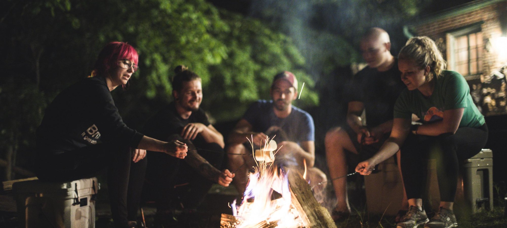 A group of five people sit around a glowing campfire at night, roasting marshmallows. Trees and a cabin are faintly visible in the background. The mood is warm and cheerful.