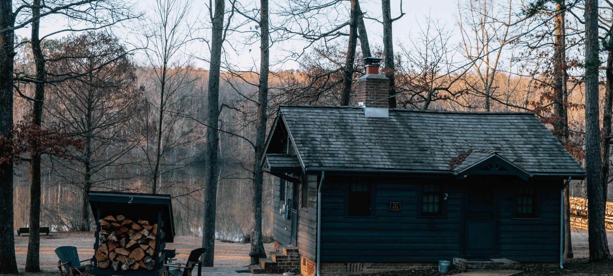 A rustic cabin with a chimney is nestled among leafless trees in a serene, wooded area. A stack of firewood is nearby, creating a cozy, tranquil atmosphere.
