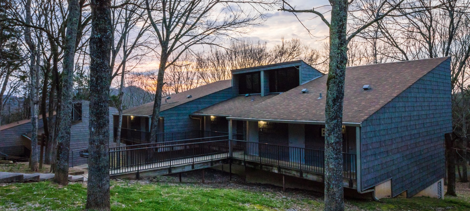 Modern building with dark wood siding and large windows surrounded by bare trees. A sunset sky casts a warm glow, creating a serene atmosphere.