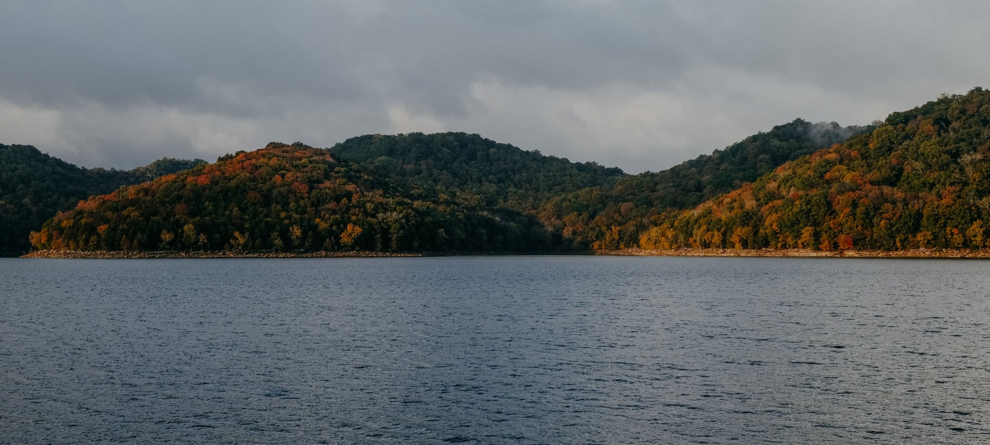 A serene lake with rippling water is in the foreground, surrounded by lush, forested hills displaying autumn colors under a cloudy sky.