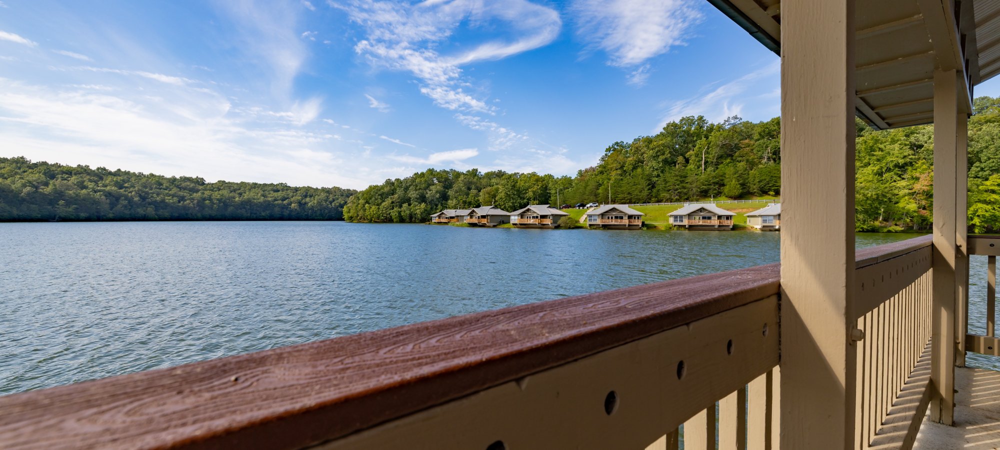 View from a shaded balcony overlooking a tranquil lake, with cozy cabins nestled along the green tree-lined shore under a clear blue sky.