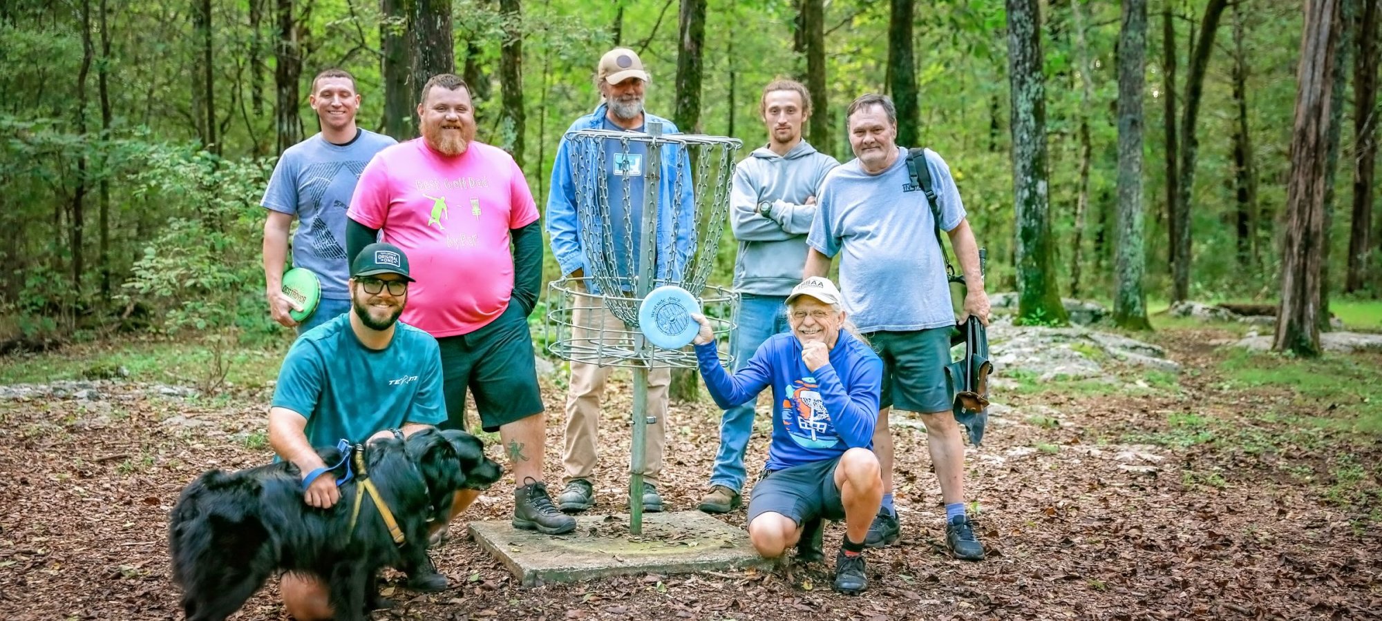 A group of seven people and a dog are gathered around a disc golf basket in a wooded area. They appear cheerful, with one person holding a disc. The setting is lush and green.