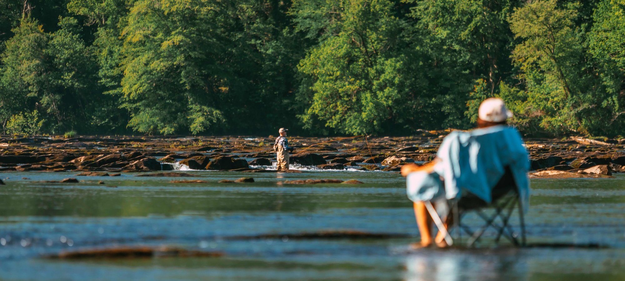 Man sitting in a chair outside by water while a man in the distance is in the water fishing