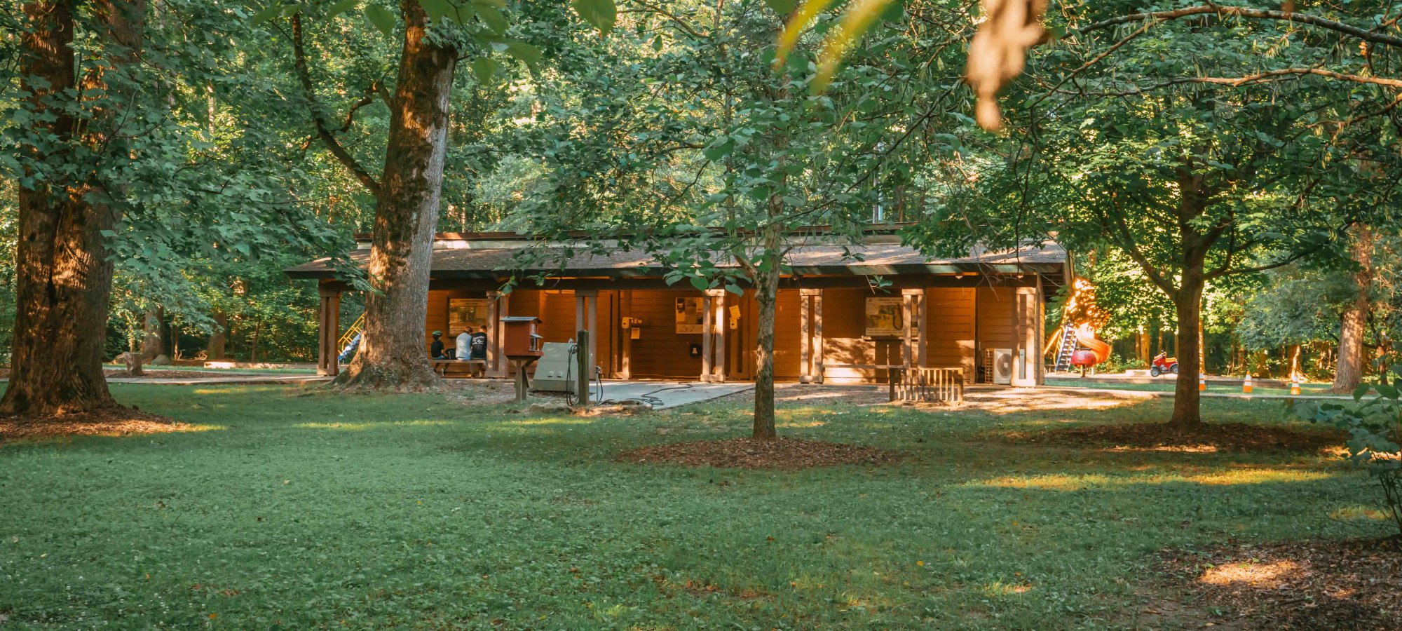 view of a flat green area with a pavilion like shelter surrounded by some trees and a playground in the background