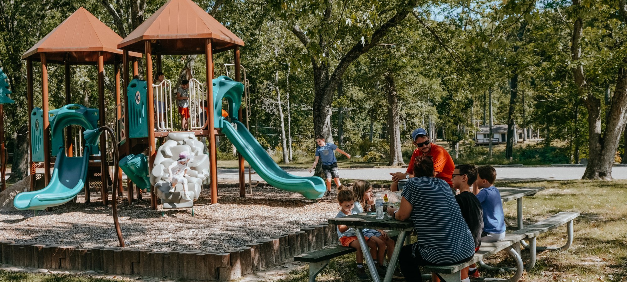 Kids play on a vibrant green playground with slides, while a family sits at a nearby picnic table. The setting is a sunny park with trees.