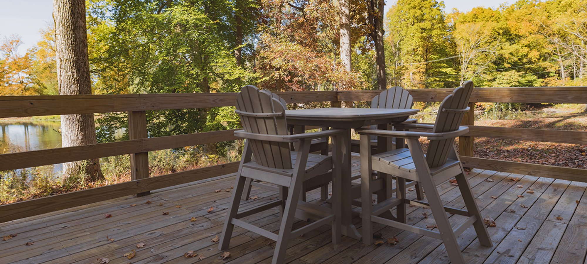 A wooden patio deck features a table and four chairs, surrounded by autumn trees with colorful leaves. The scene is peaceful and sunlit.