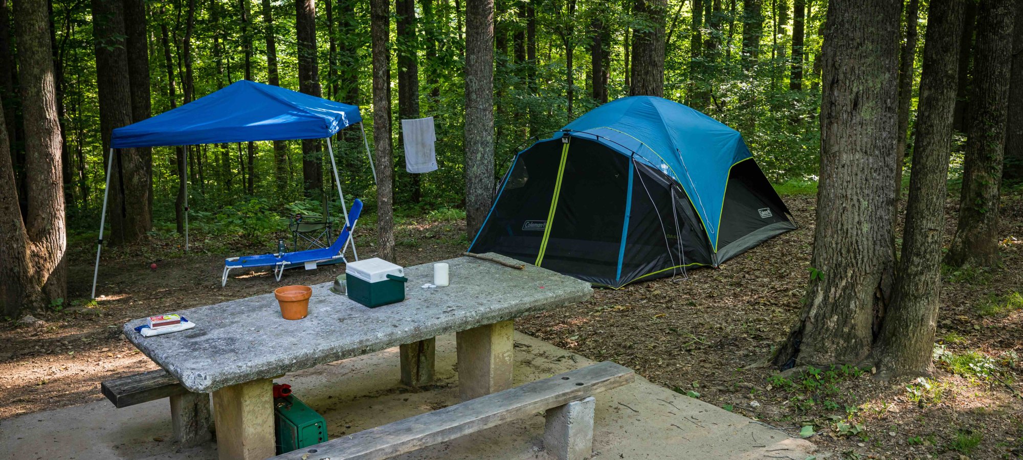 view of camping area with a picnic table with containers on top, a tent and a lounge chair surrounded by trees