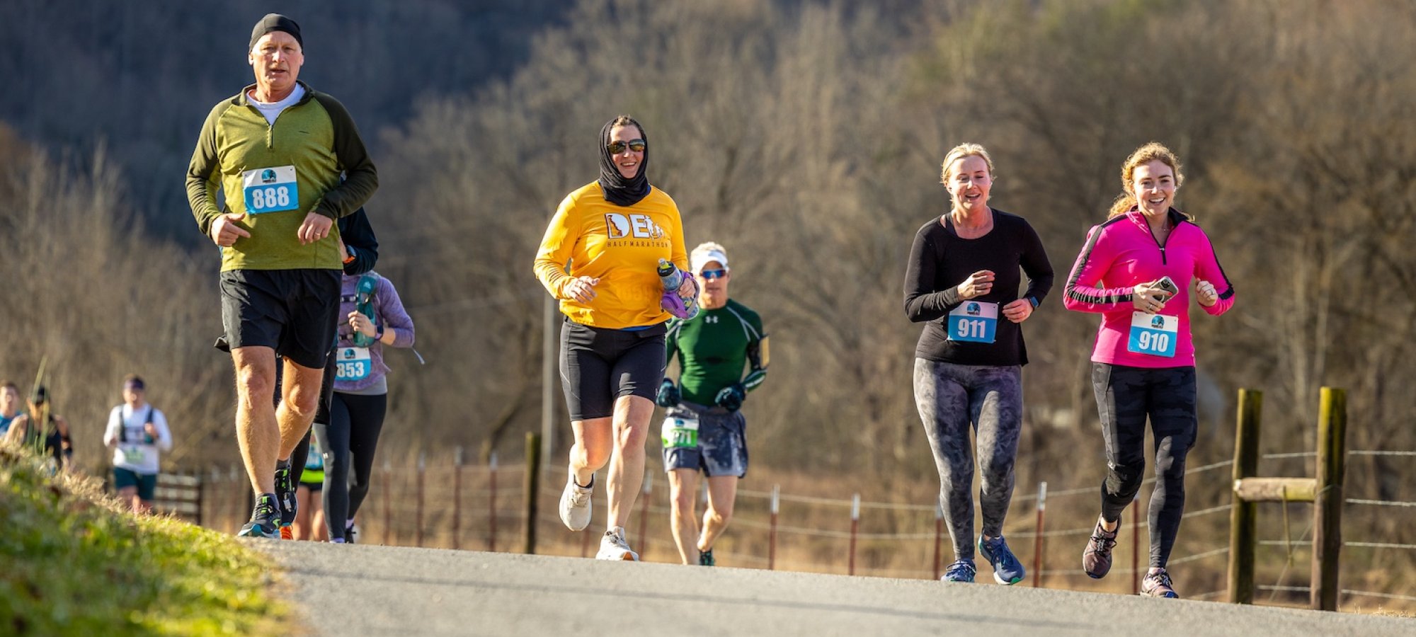 A group of runners on a rural path, wearing colorful athletic gear. The scene is bright and lively, with trees and blue skies in the background.