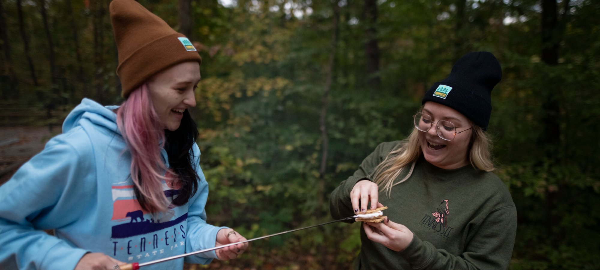Two people in beanies enjoy making s'mores over a fire in a forest, sharing laughter and a sense of camaraderie amid autumn foliage.