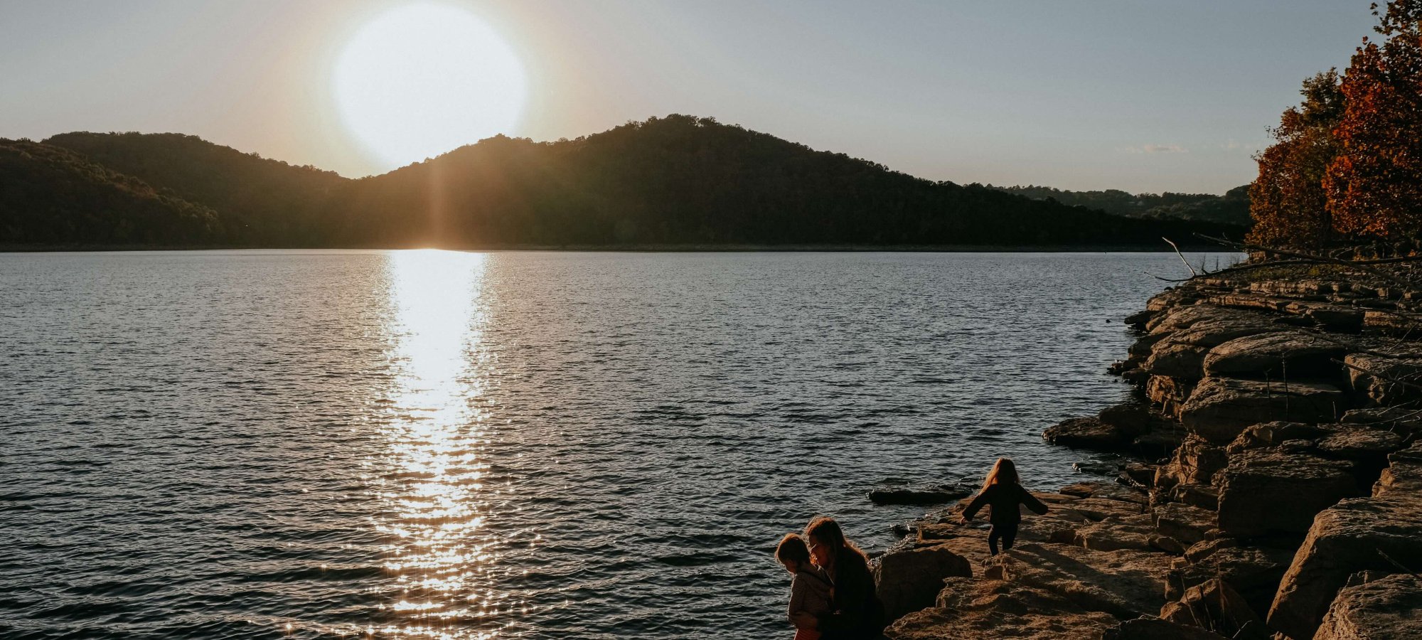 Sunset over a lake with people sitting on rocky shore.