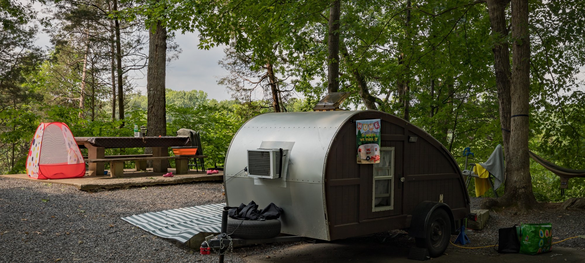 A cozy campsite with a small vintage trailer and a red tent surrounded by lush trees. A picnic table and hammock suggest a peaceful outdoor setting.