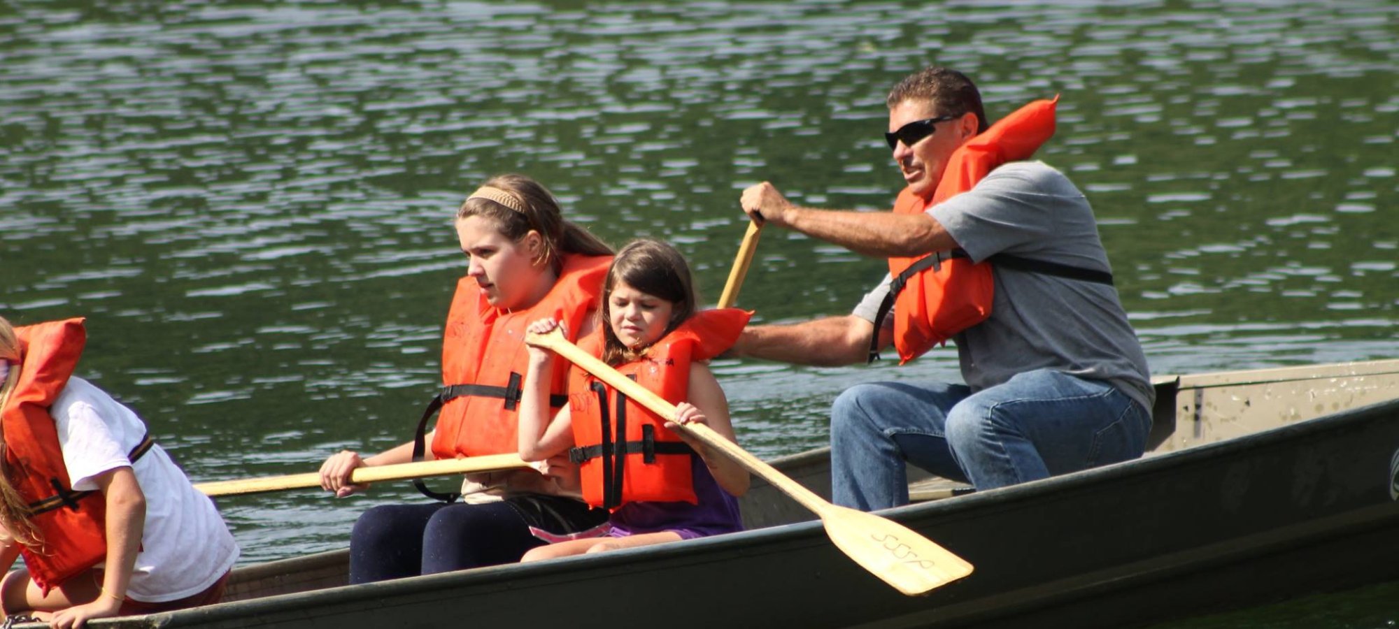 Family with life jackets paddling a canoe on a calm lake.