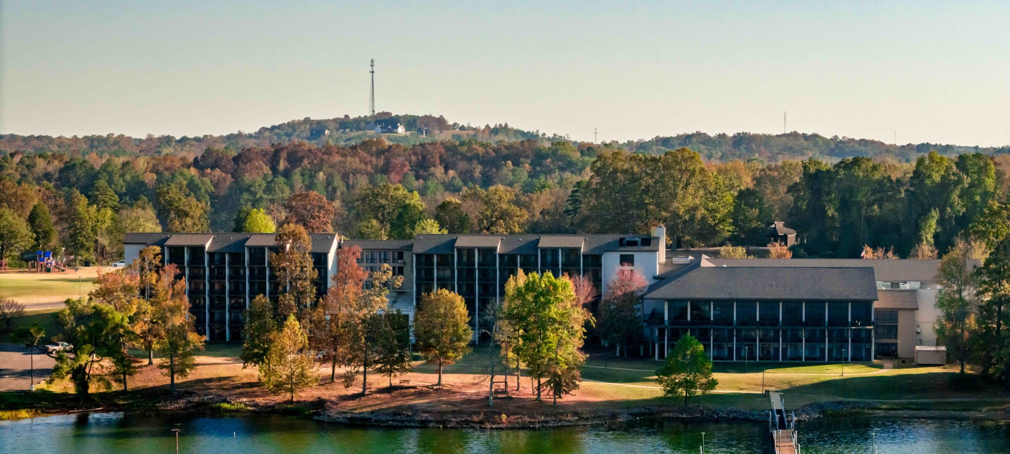 Lakeside resort buildings surrounded by lush trees with a hill in the background.