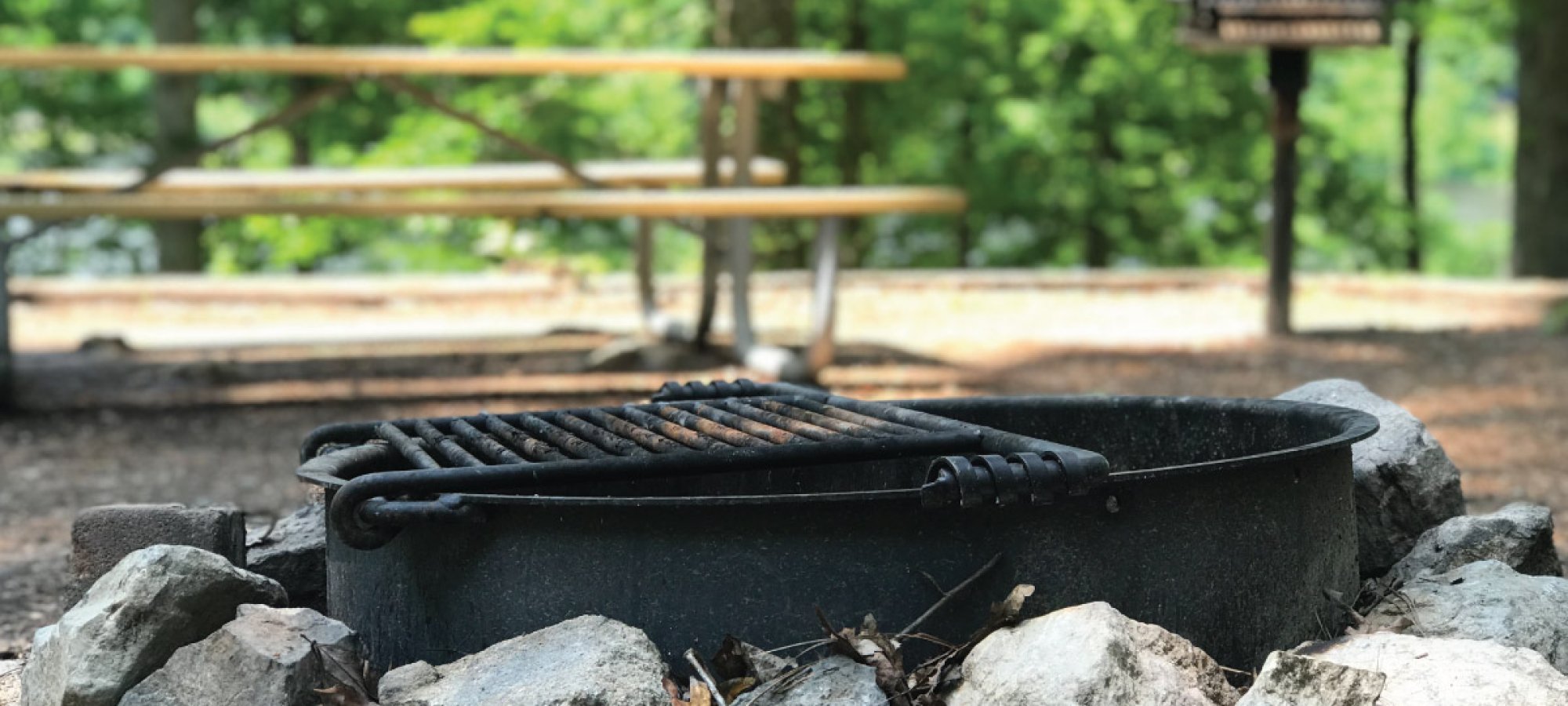 Outdoor grill surrounded by stones with picnic tables in background.