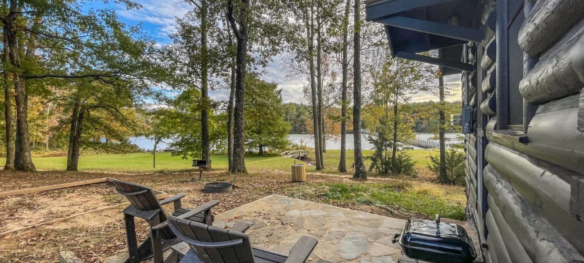 Patio with chairs beside a cabin overlooking a serene lake surrounded by trees.