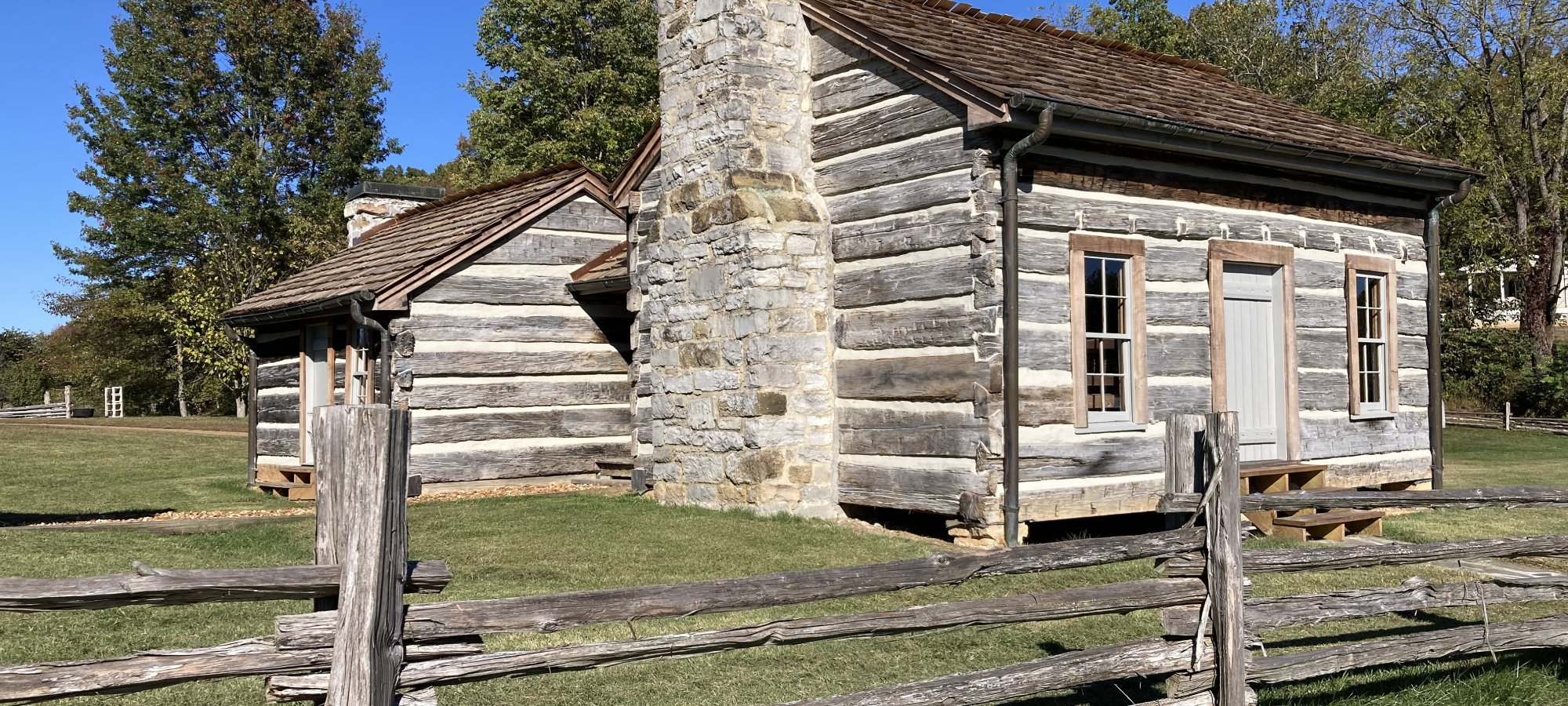 A rustic wooden cabin with a stone chimney and weathered appearance sits behind a wooden fence. Surrounded by grass and trees under a clear blue sky.