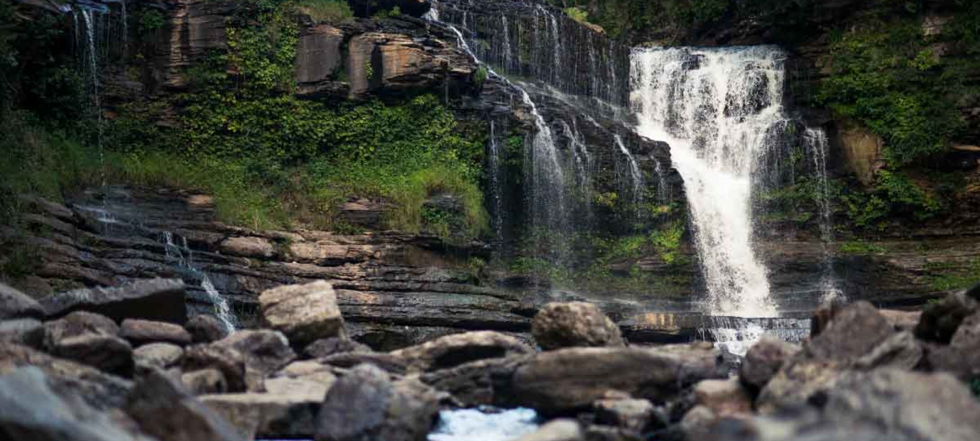 Waterfall cascading over rocky cliff into a boulder-strewn river.
