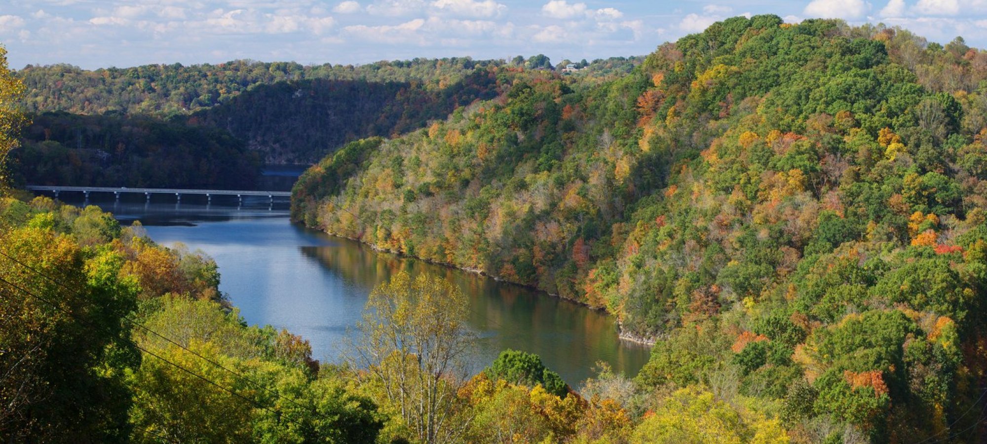 Scenic view of a river winding through lush, tree-covered hills showcasing autumn colors. Fluffy clouds dot a bright blue sky, evoking tranquility.