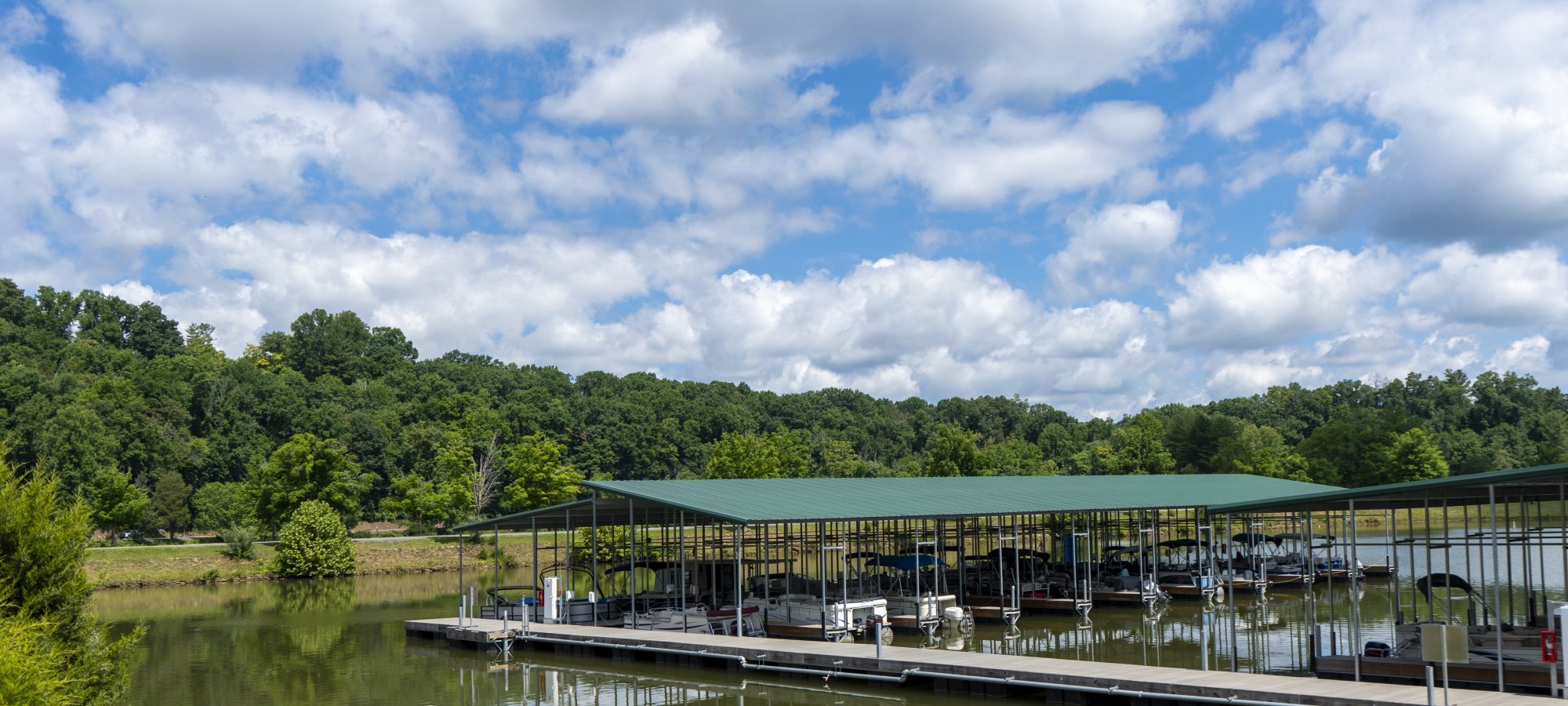 A serene lakeside view with a large covered boat dock, surrounded by lush, green trees. The sky is blue with scattered white clouds, reflecting in the calm water.