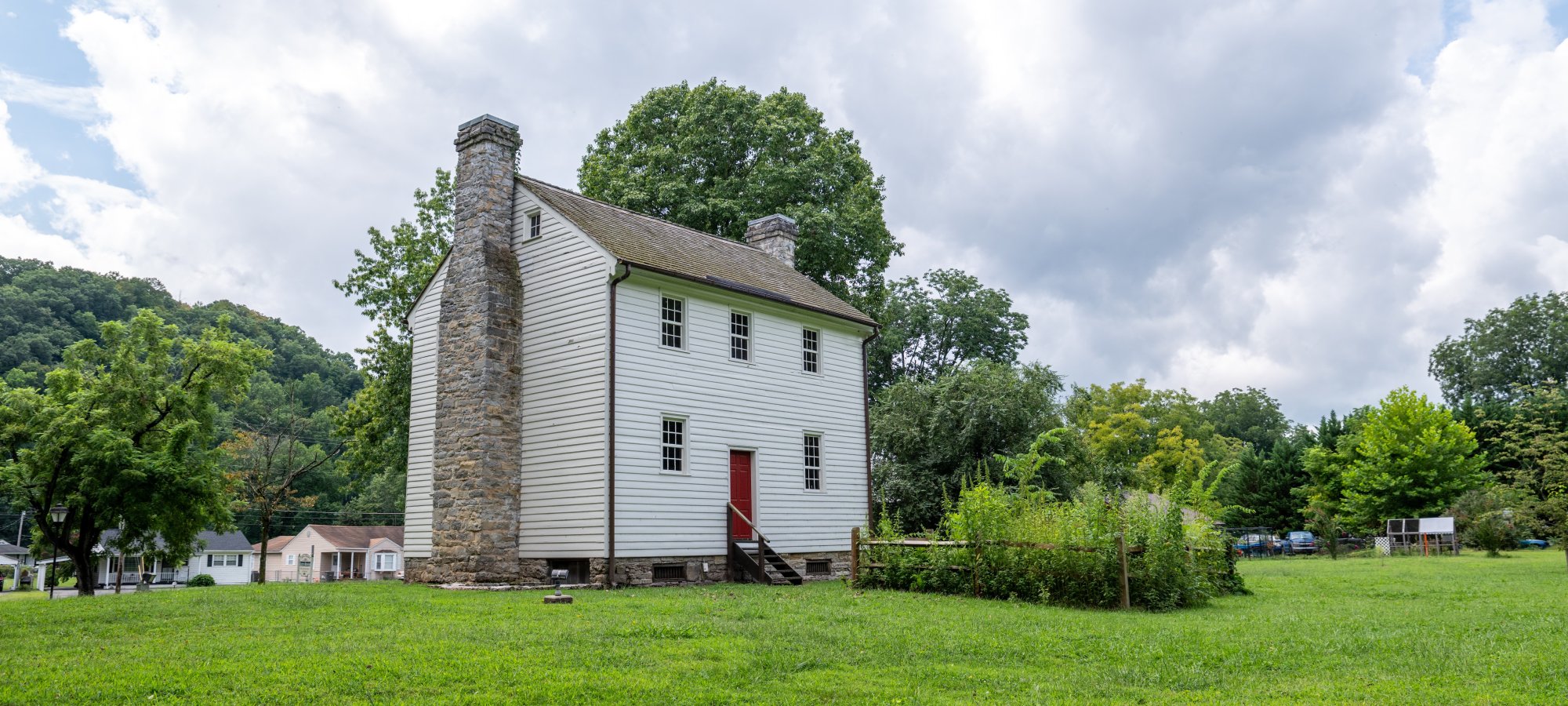 Historic stone house with red door, surrounded by lush lawn and trees under cloudy sky.