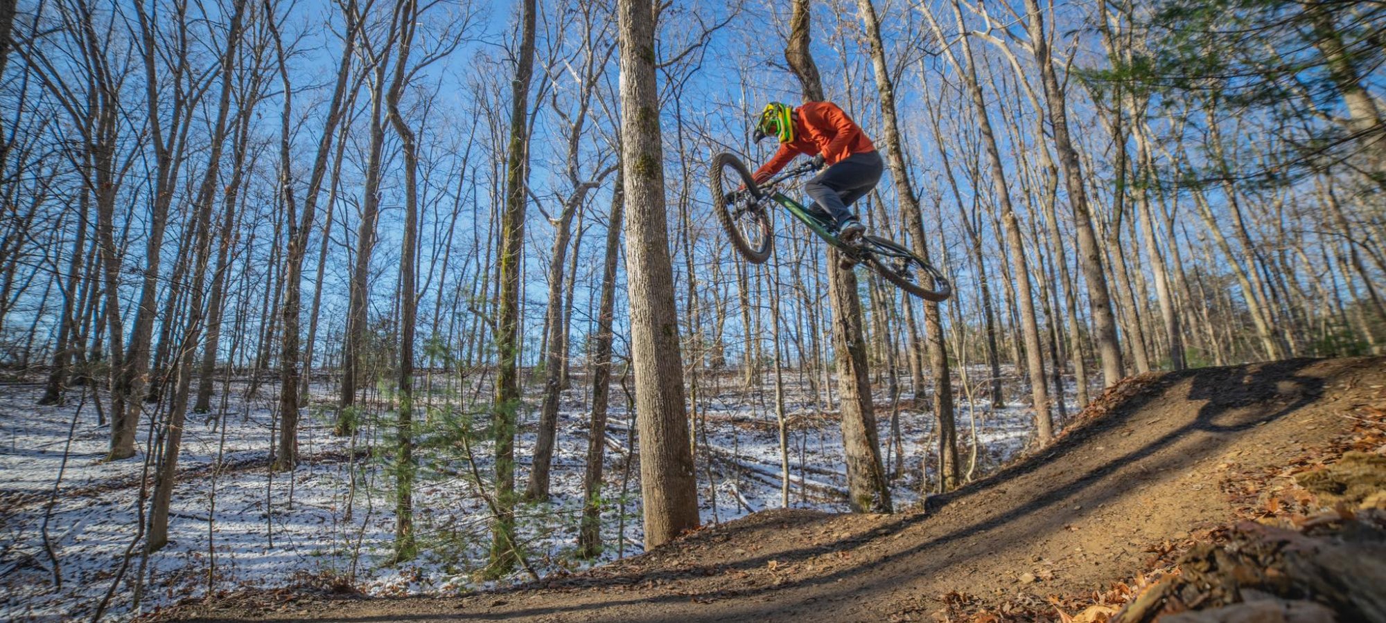 Mountain biker mid-air on a forest trail with snow patches.