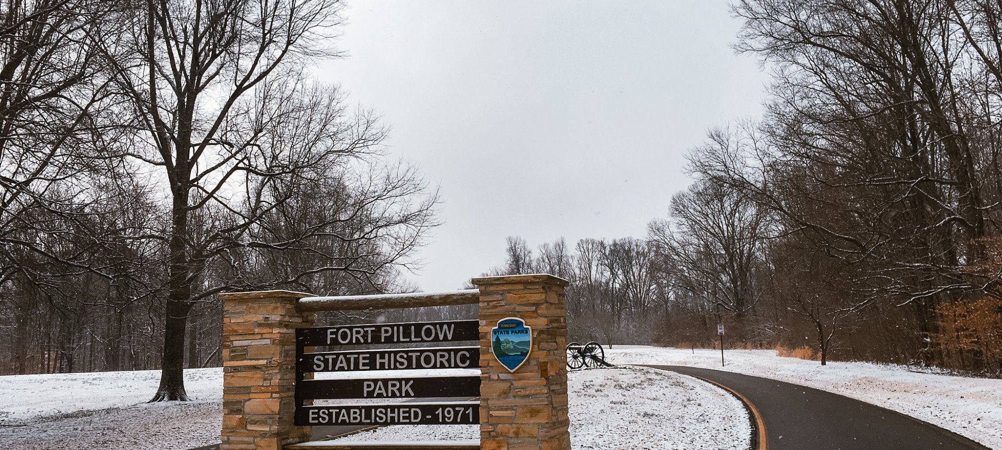 Entrance sign in snowy park with bare trees and a winding road.