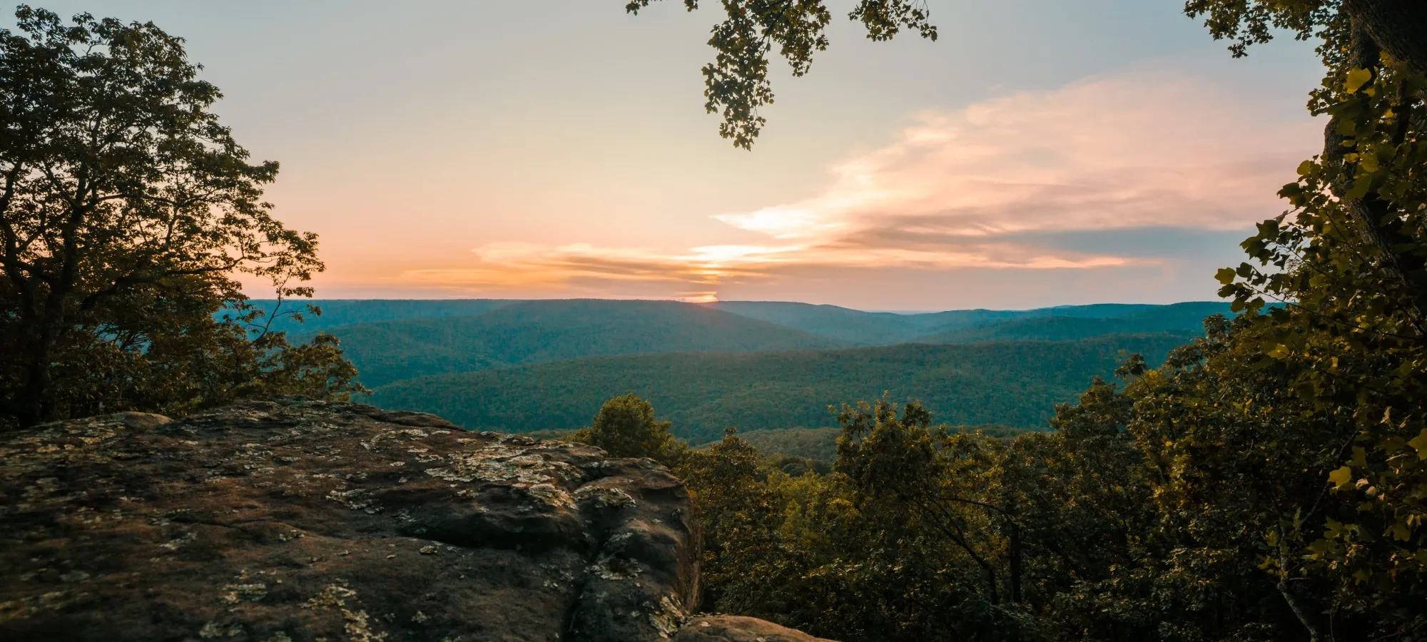 Sunset over a forested valley viewed from a rocky overlook.
