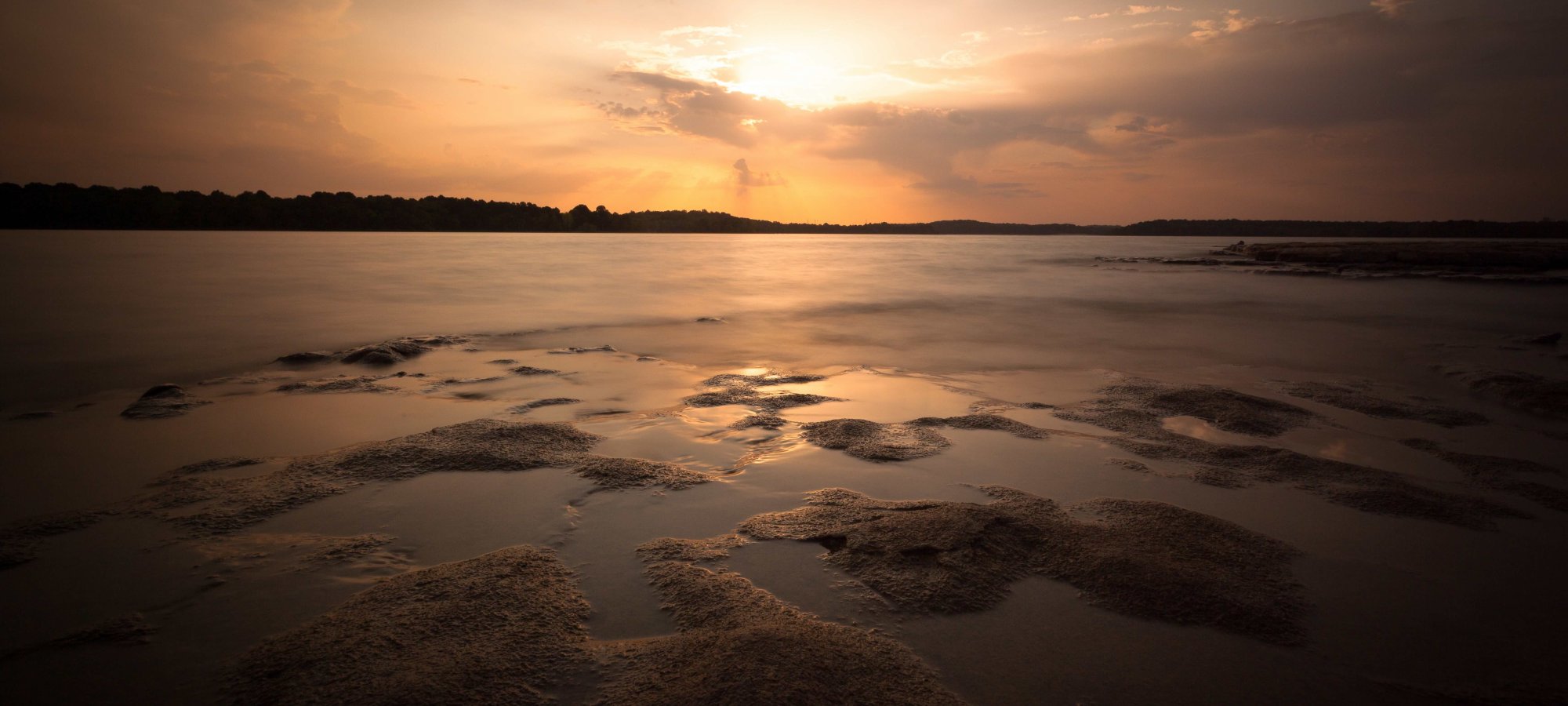 Sunset over a tranquil river with sandy shores and cloudy sky.