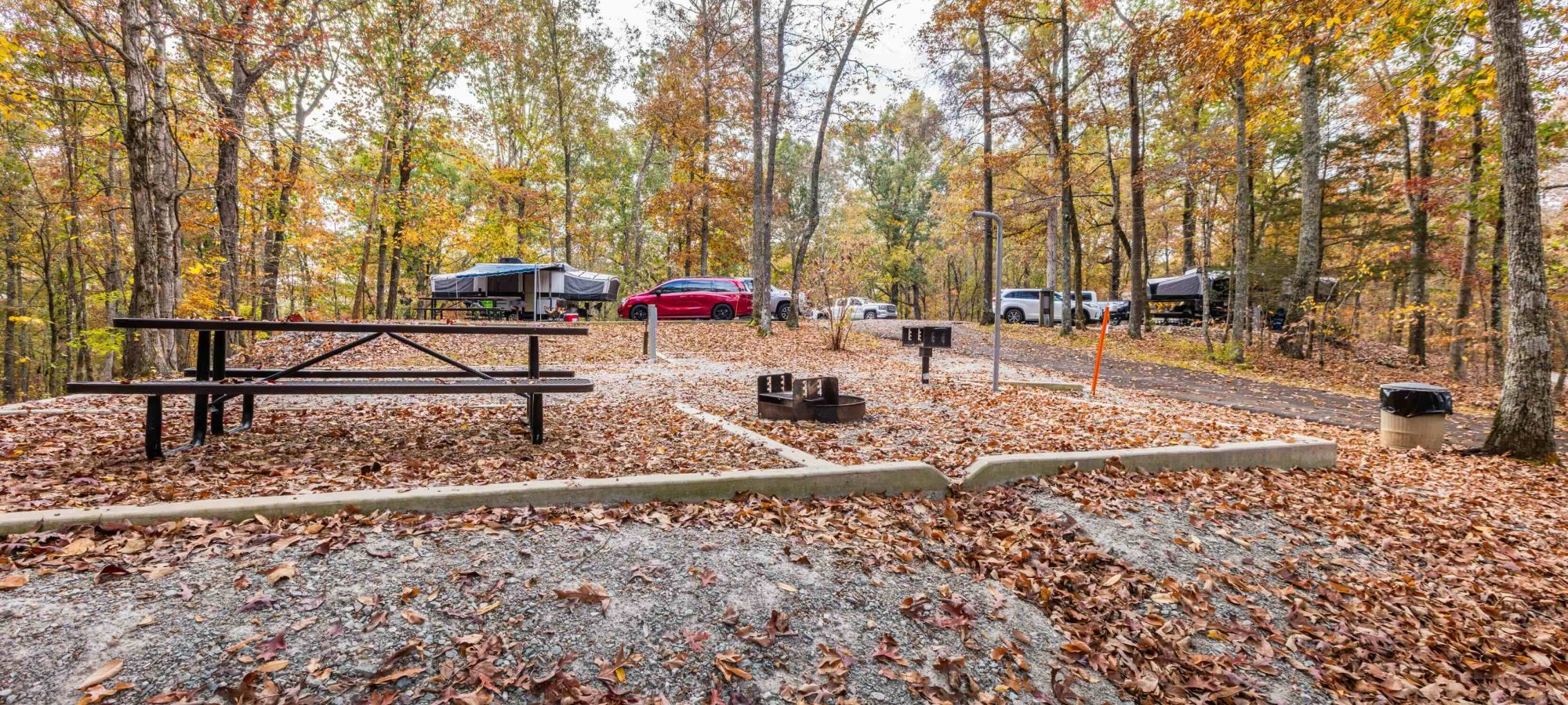 Autumn campground scene with tents, vehicles, and a picnic area surrounded by trees.