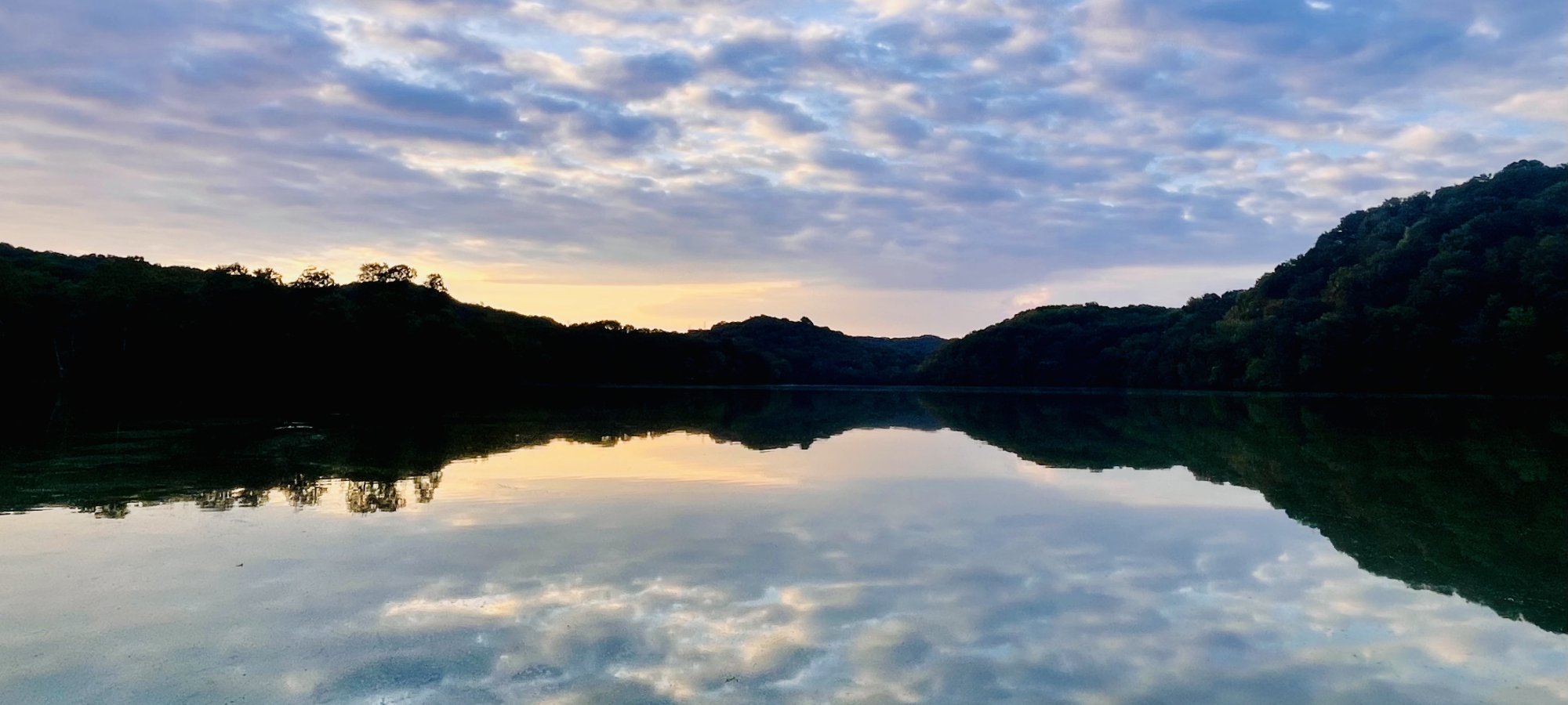 Sunset over a calm lake with reflections of clouds and tree-lined hills.