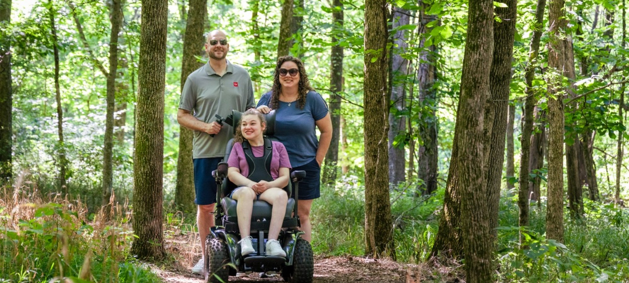 Child in a motorized wheelchair exploring the forest with two adults.