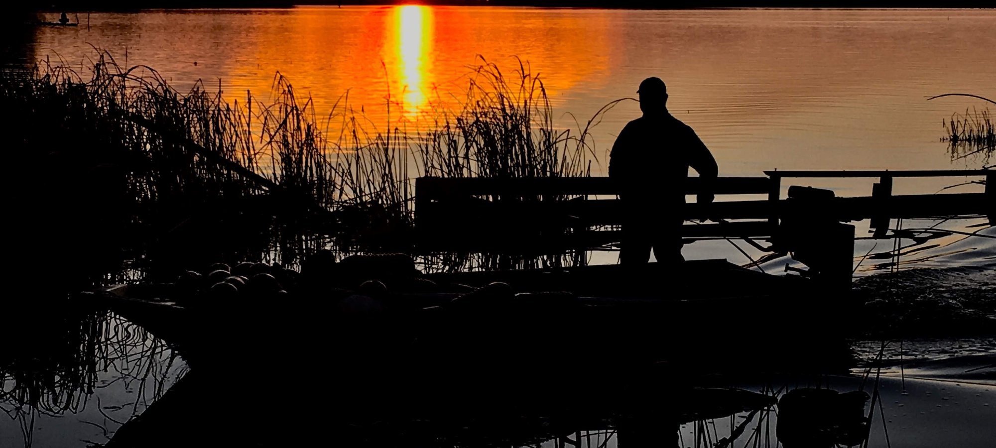 Silhouette of a person fishing on a lake dock at sunset.