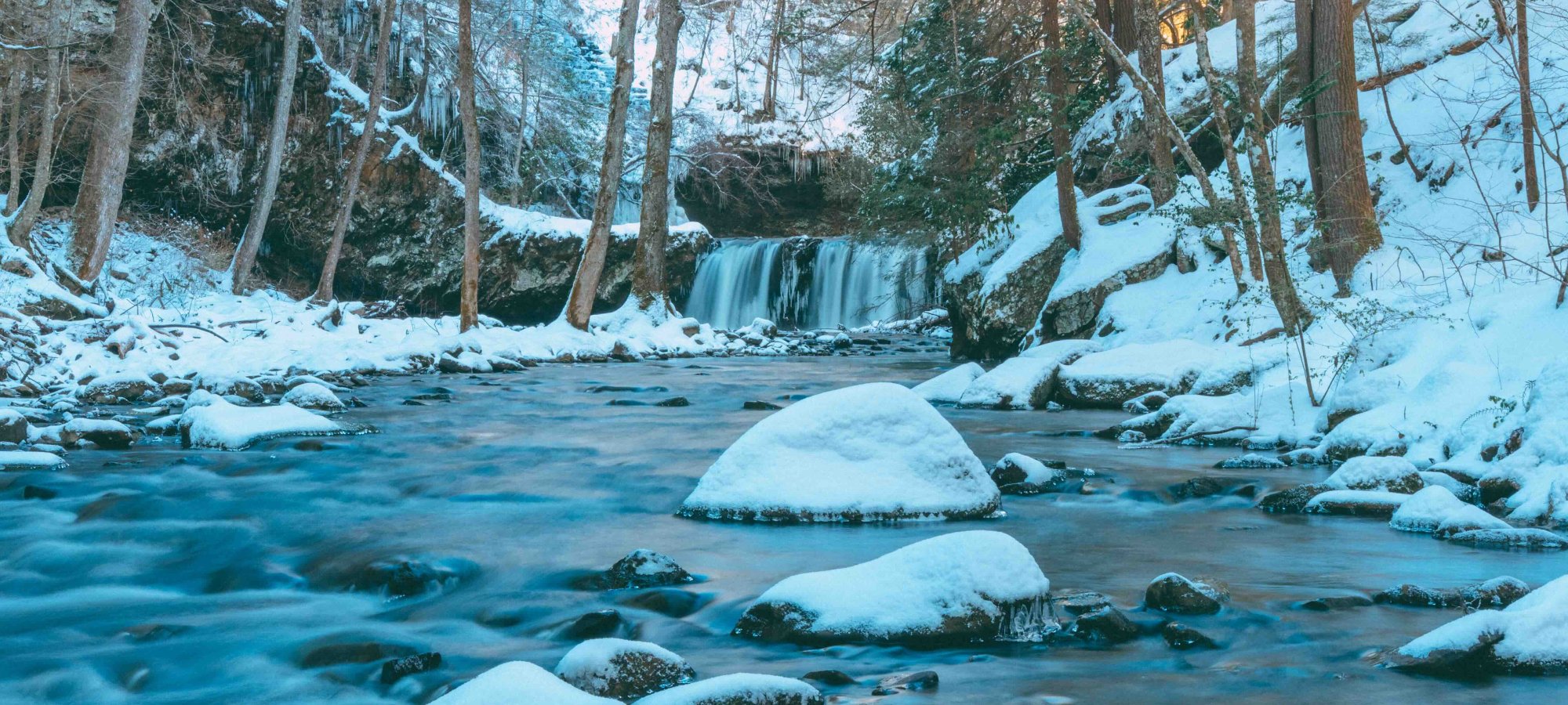 Snow-covered river with rocks and a small waterfall in a forest.
