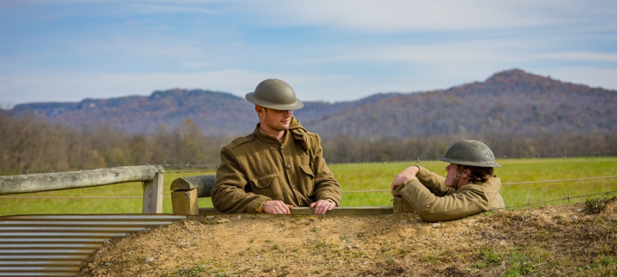 Two men dressed as World War One soldiers stand face to face in a trench in a bright green field with a hazy view of mountains behind them. 