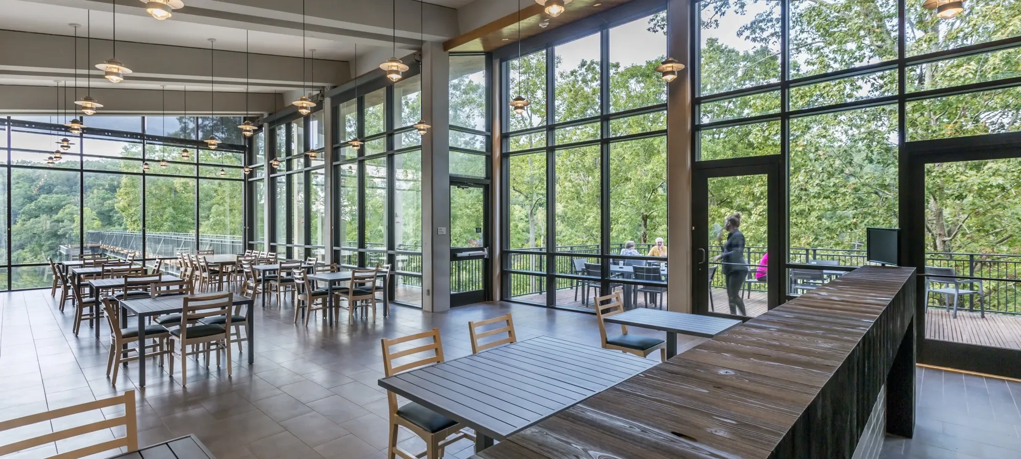 The interior of a restaurant with large floor to ceiling windows. There are tables and chairs inside and you can see outside that there is a patio with more seating options.