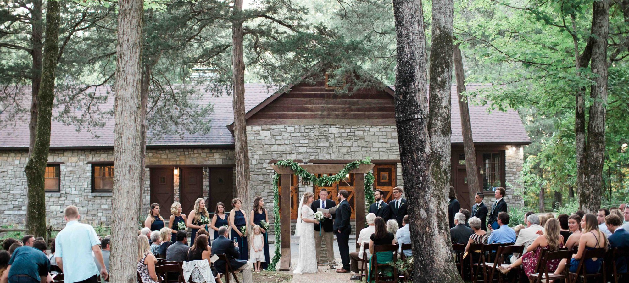Outdoor wedding ceremony in a forest, guests seated along a path leading to a wooden cabin.