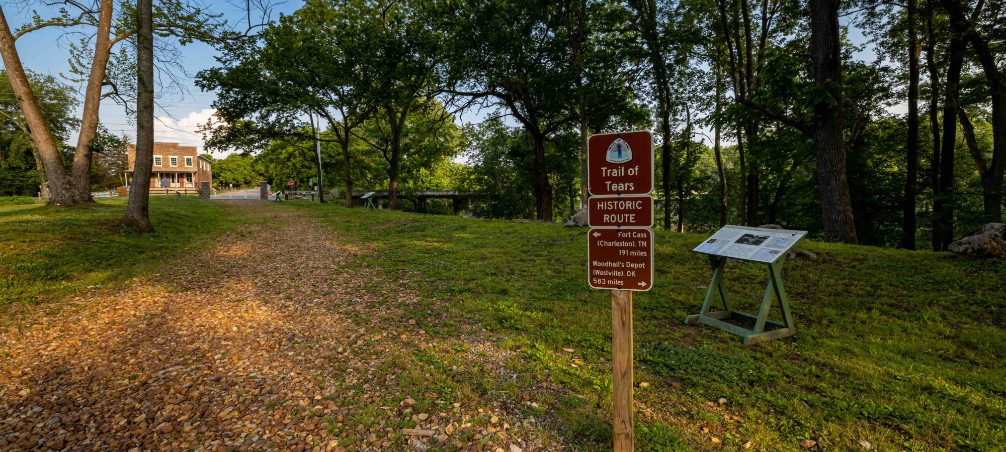 Trailhead with informational signs and a path leading to a distant house surrounded by trees.