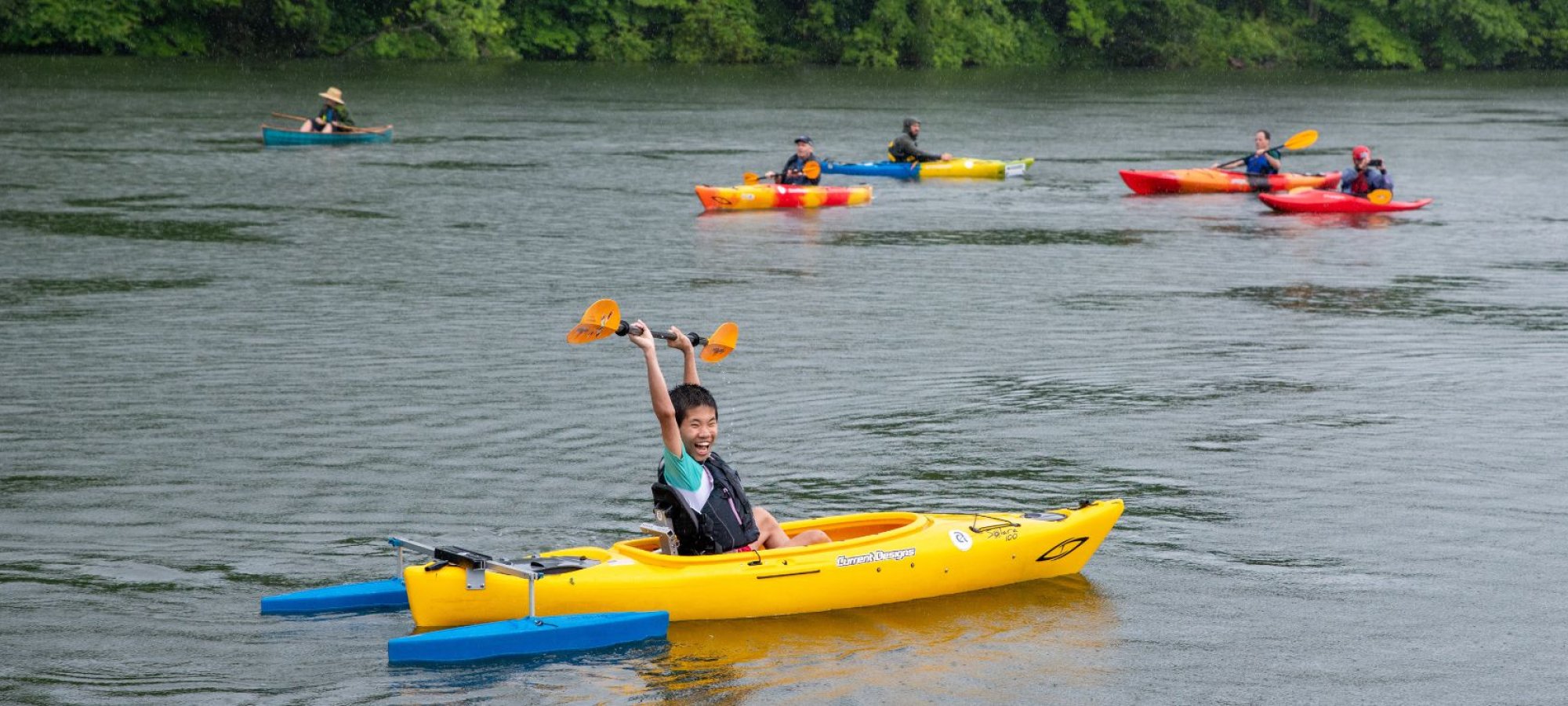 A child in a yellow kayak smiling and holding a paddle above their head while others paddle in the distance on a peaceful river.