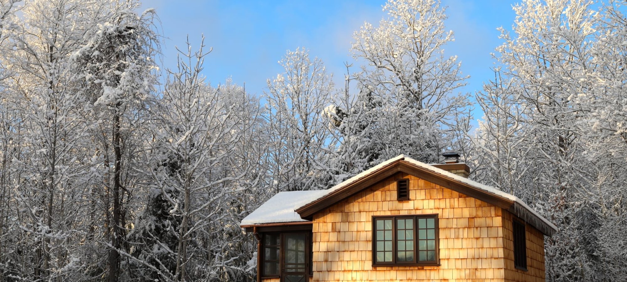 A collage showing a rustic cabin interior with wood walls, exposed beams, large windows, and a stone fireplace, alongside an exterior view of a small wood‑and‑stone cabin under a bright sky with scattered clouds.