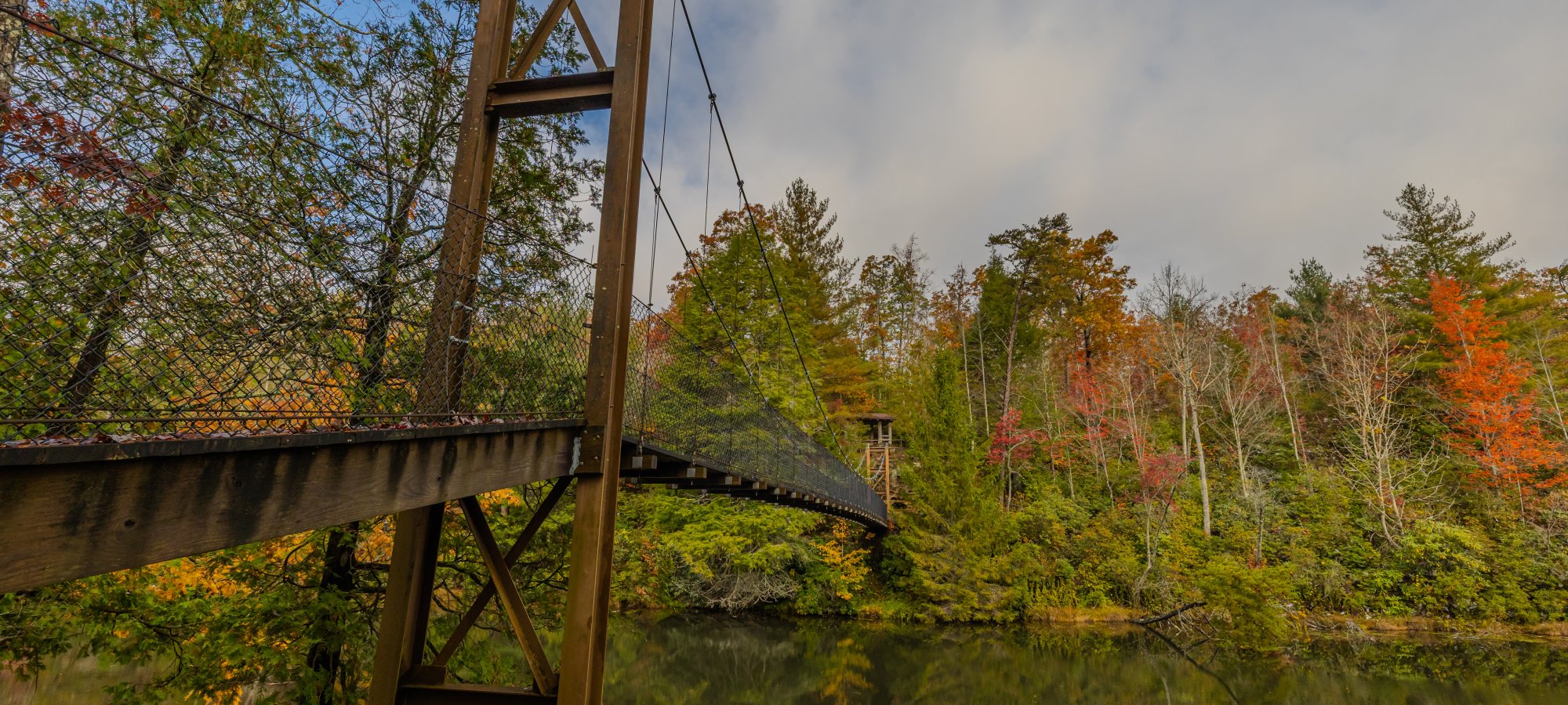 Suspension bridge over a calm lake surrounded by colorful autumn trees under a partly cloudy sky.