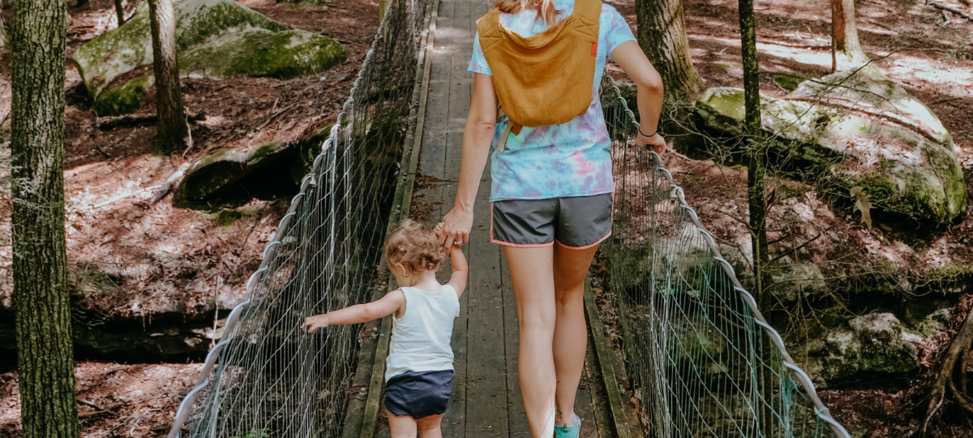 A mother and small child hold hands while crossing a suspension bridge in the woods.