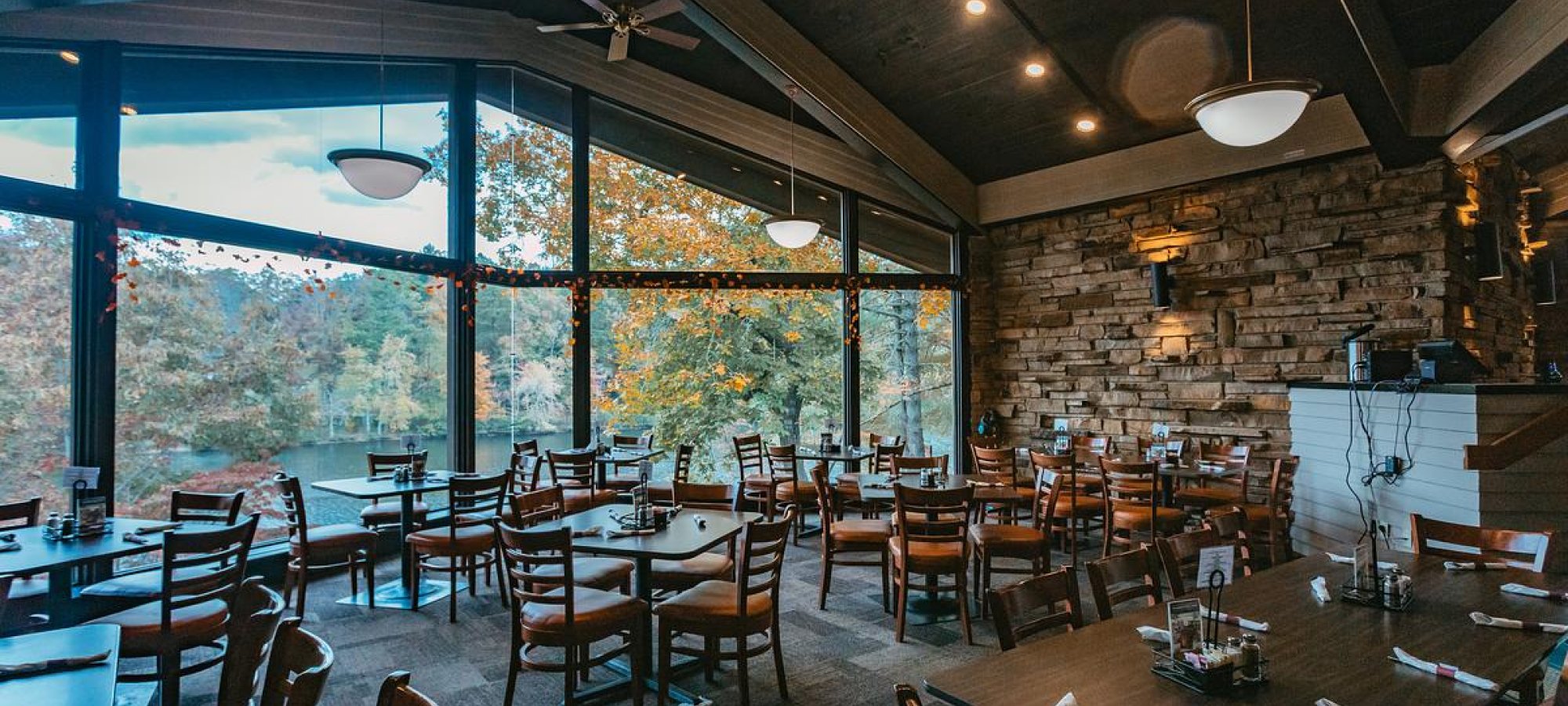 Cozy restaurant interior with large windows, wooden tables, and autumn view outside.