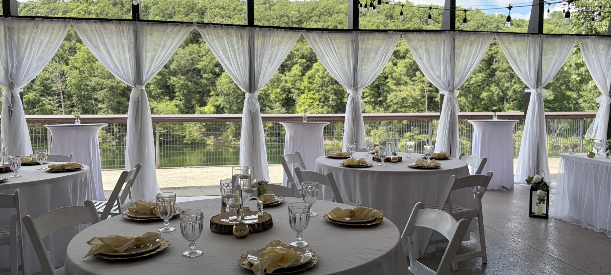 Wedding reception setup with white-draped tables, sheer curtains, string lights, and a lakeside view surrounded by trees.