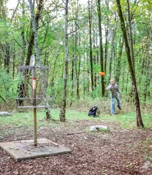 Person playing disc golf in a forest, throwing an orange disc toward a metal basket.
